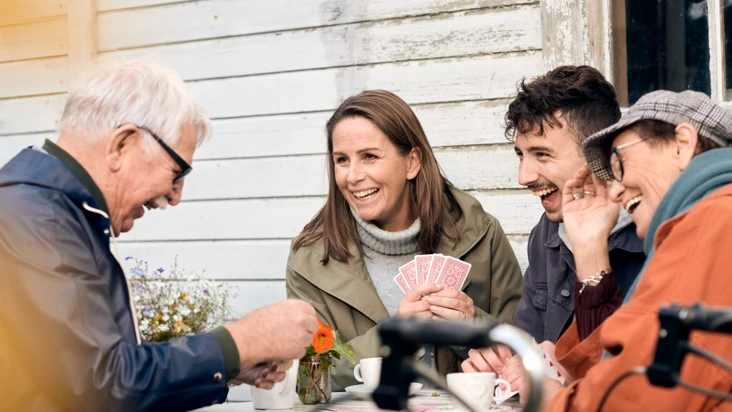 Två pensionärer och två yngre personal i hemtjänsten spelar kort och skrattar tillsammans.