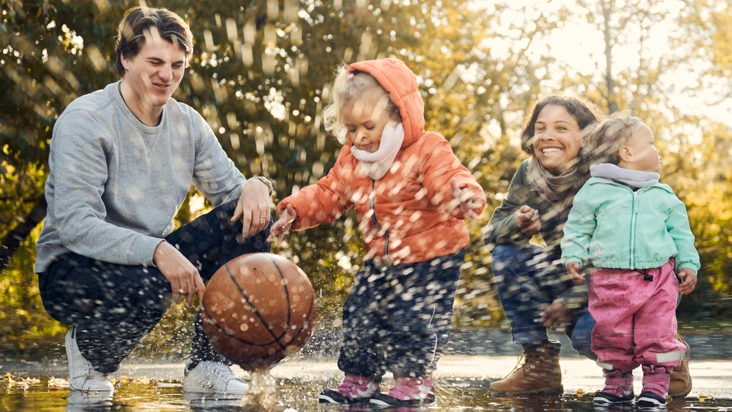 Ett litet barn släpper en boll i en vattenpöl så det skvätter vatten på förskolepersonalen som skrattar.